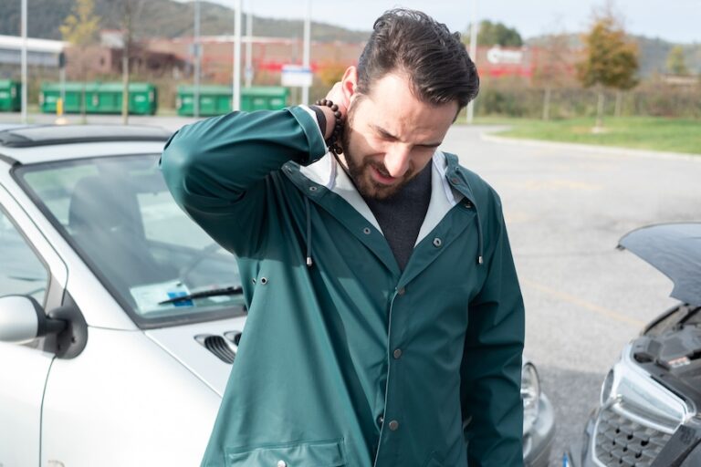 A white grown male, in a green jacket, is having neck pain and is experiencing whiplash symptoms after a head-on car collision.