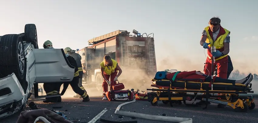 Paramedics and firefighters work urgently around an overturned vehicle on a smoky road with a fire truck in the background and a stretcher nearby depicting the chaotic emergency response to a serious crash involving life-threatening car accident injuries in Metro Atlanta.