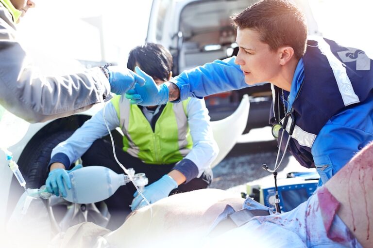 Two paramedics in blue gloves and safety vests administer emergency oxygen to a bloodied accident victim on the ground beside a vehicle representing the critical on-scene medical treatment required for severe car accident injuries before transport to a hospital in Metro Atlanta.