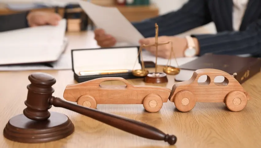 A wooden judge's gavel and two wooden toy cars staged in a rear-end collision sit on a desk in front of an attorney reviewing documents beside scales of justice representing the legal process of pursuing compensation for car accident injuries at Atlanta Accident Lawyers.