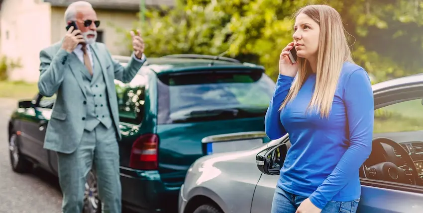 A young woman in a blue long-sleeve shirt and an older man in a gray three-piece suit stand beside their damaged vehicles on a tree-lined street both on separate phone calls with Atlanta Accident Lawyers after a rear-end collision in a situation where police never showed up to take a report in Metro Atlanta.