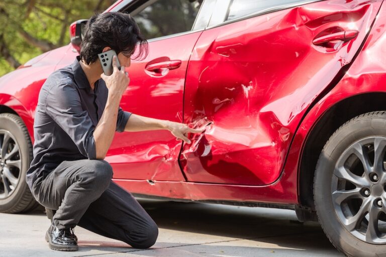 A dark-haired man in a navy shirt crouches beside a red car pointing at a deep dent and crumpled panel on the driver's side door while talking on his phone documenting the collision damage himself after police never showed up to the accident scene in Metro Atlanta.