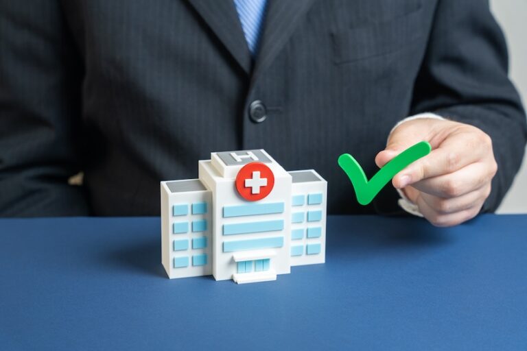 A lawyer- from Atlanta Accident Lawyers- in a dark pinstriped business suit sits at a blue surface and holds a bright green three-dimensional checkmark symbol in their right hand beside a detailed miniature model of a white hospital building with blue-tinted windows- a red circular emblem with a white cross on its facade- and a large letter H on the upper portion of the structure — representing the formal approval process of selecting an approved doctor or medical facility recognized by an insurance provider for treatment following an injury in Metro Atlanta.