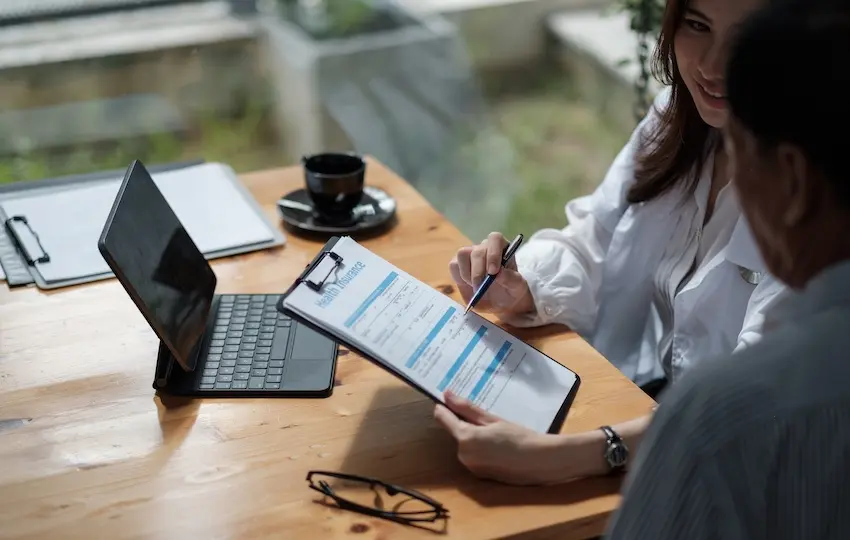 A smiling female insurance agent points to a car insurance form on a clipboard while reviewing an insurance policy with a client at a wooden desk beside a laptop and coffee cup in Atlanta.