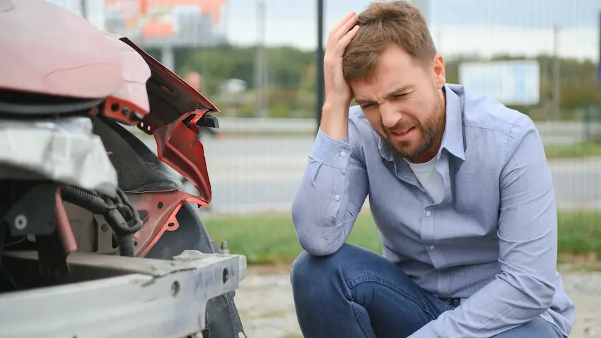 A distressed man crouches beside the severely damaged rear end of a red vehicle while holding his head in his hands representing the overwhelming stress of dealing with an insurance policy claim after a serious collision in Atlanta.