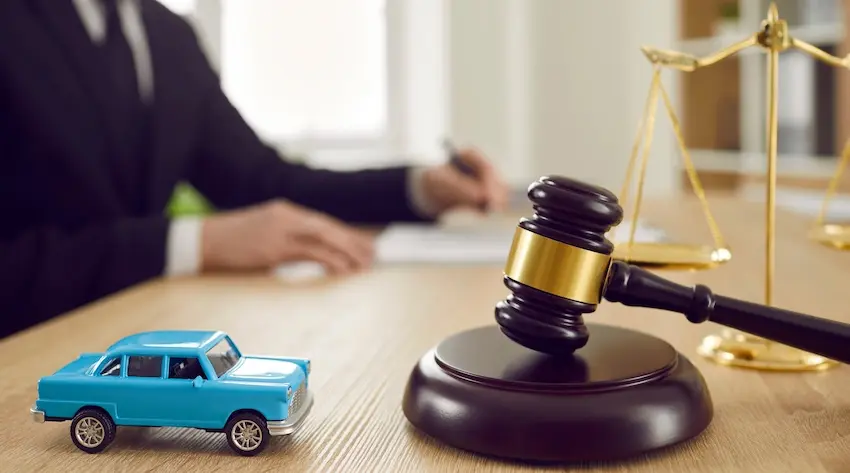 A blue toy car and a dark wooden judge's gavel on a sound block sit on a wooden desk at Atlanta Accident Lawyers in the foreground while a person in a black suit writes on documents in the background beside a gold balance scale representing the legal proceedings where dashcam video footage is submitted as evidence in a car accident case.