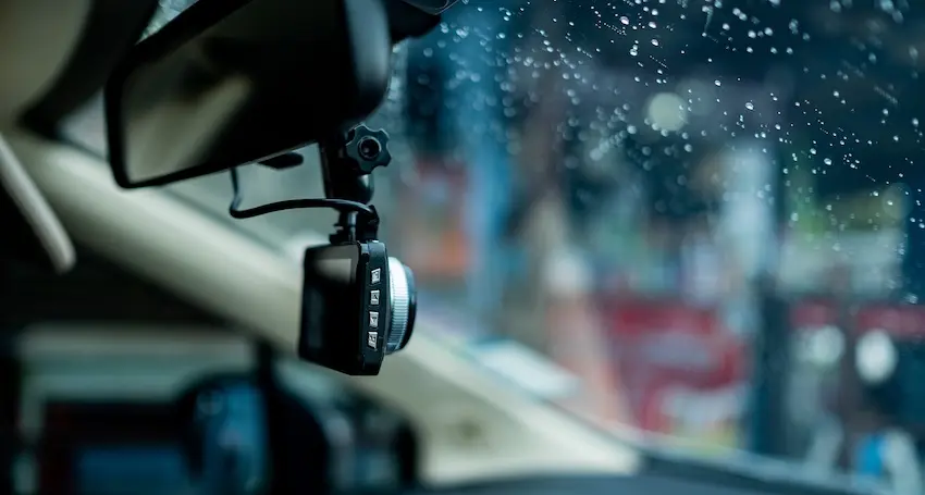 A compact black dashboard camera with a wide-angle lens mounted near the rearview mirror of a vehicle records through a rain-covered windshield on a wet city street with blurred traffic and stormy conditions in the background showing how dashcam video continues recording even in adverse weather when accidents are most likely to occur.