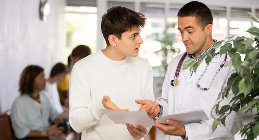 A young male patient with short dark hair wearing a white crew-neck sweater gestures with an open hand in a frustrated manner while holding a document at his side- standing beside a male doctor in a white lab coat with a dark purple stethoscope draped around his neck who is pointing at a separate sheet of paper and explaining something- both standing near a large green potted leafy plant in a bright medical office lobby with blurred figures seated in the background — depicting the kind of disagreement a patient might have when navigating care with an approved doctor under an insurance plan in Metro Atlanta.