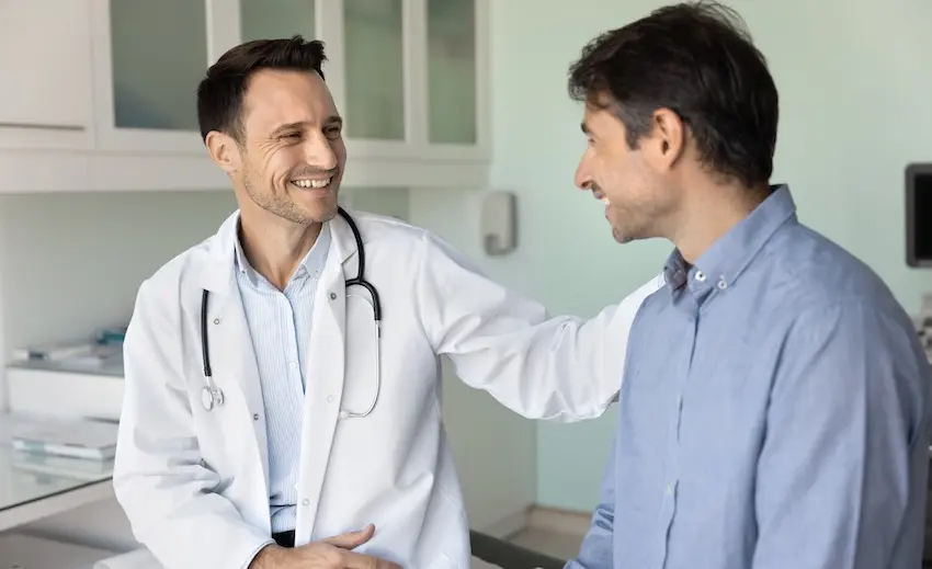 A smiling male doctor with short dark hair and light stubble wearing a white lab coat with a black stethoscope around his neck extends his arm warmly to place a reassuring hand on the shoulder of a male patient in a light blue button-down shirt who is facing him with a slight smile- both standing in a clean modern clinical examination room with white cabinetry- a wall-mounted dispenser- and soft teal walls visible in the background — illustrating the positive and supportive experience patients can expect when receiving care from an approved doctor- with assisstance from Atlanta Accident Lawyers.