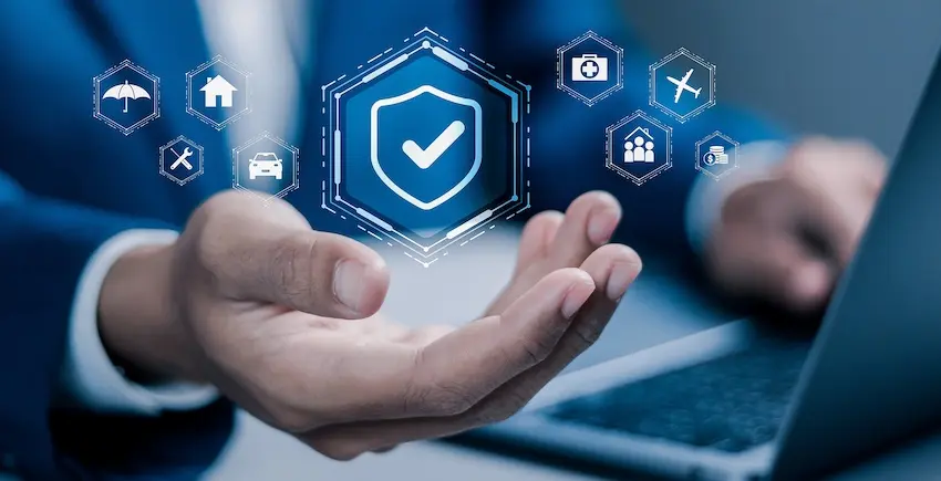 A person in a blue business suit holds one open palm upward beneath a glowing three-dimensional blue hexagonal shield icon with a white checkmark at its center surrounded by six smaller floating hexagonal icons representing different categories of insurance coverage including an umbrella for general coverage with a blurred laptop visible in the background — a digital concept image representing the broad protection offered when working with an approved doctor through a comprehensive insurance company network in Metro Atlanta.