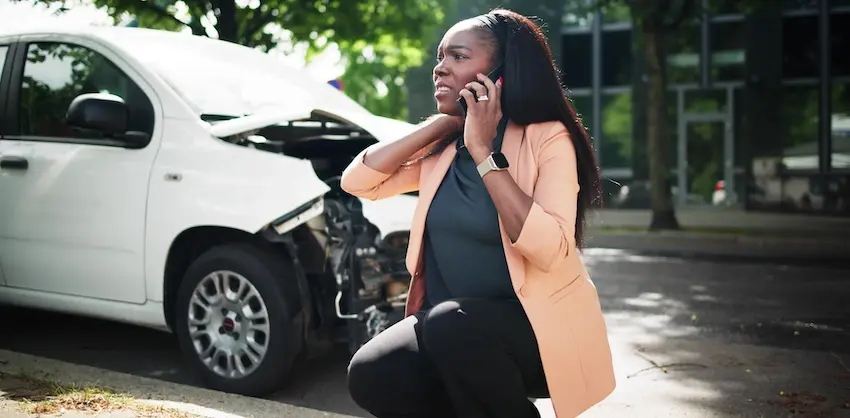 A woman in a peach blazer crouches on a curb while talking on her phone to Atlanta Accident Lawyers in Metro Atlanta and clutching her neck in pain with a white van behind her showing severe front-end damage with the hood crumpled open highlighting the importance of dashcam video in documenting the full impact of a crash and supporting a victim's injury claim.