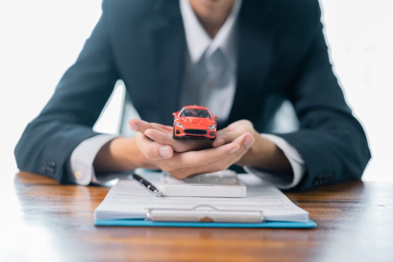 A suited lawyer from Atlanta Accident Lawyers cups both hands around a small orange model car over a clipboard with documents and a pen on a wooden desk symbolizing the protection and coverage provided by a comprehensive auto insurance policy.