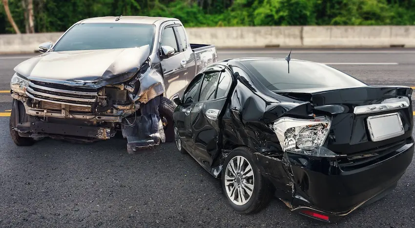 A silver pickup truck with a destroyed front end sits pushed against a black sedan with a heavily crumpled rear quarter panel on a highway showing the severity of a rear-end collision that can leave victims requiring future medical treatments for years after the crash in Metro Atlanta.