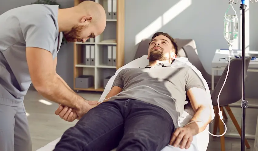 A bald bearded male nurse in gray scrubs inserts an IV line into the arm of a bearded man in a gray shirt reclining in a treatment chair with an IV drip bag on a stand beside him in a sunlit clinical room depicting the kind of ongoing infusion therapy and future medical treatments that accident victims may require as part of an extended recovery plan with assistance from Atlanta Accident Lawyers.