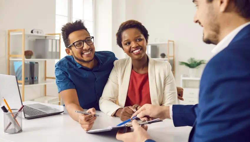 A smiling man with glasses in a blue shirt and a woman in a red top and cream blazer lean forward together at a bright office desk engaging with an attorney from Atlanta Accident Lawyers in a dark blue suit who holds a clipboard representing accident victims working with a legal professional to resolve a case where the other party disputes fault for the collision.