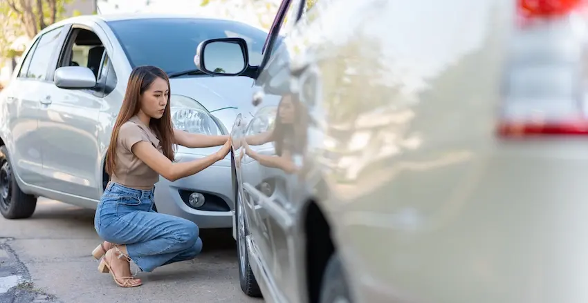 A young woman with long straight hair in a beige top and blue jeans crouches down between two cars on a roadside to closely examine the point of contact and damage where the vehicles collided illustrating the on-scene evidence gathering that becomes critical when an at-fault driver disputes fault after a crash in Metro Atlanta.