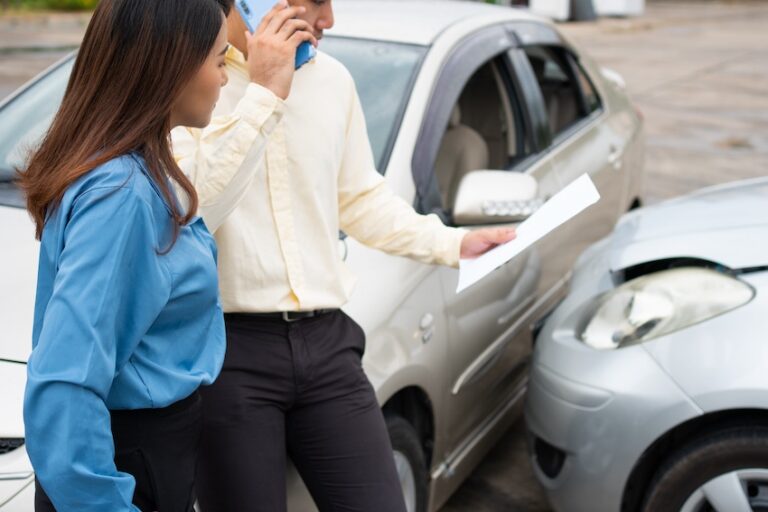 A woman in a blue shirt and a man in a yellow shirt stand beside a silver car with front-end damage as the man talks on his phone while holding out a document depicting the roadside exchange of insurance and contact information between drivers in the immediate aftermath of an accident where one party disputes fault for the collision in Metro Atlanta and could use legal support from Atlanta Accident Lawyers.